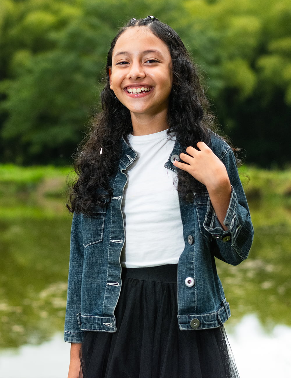 A young girl in a denim jacket and skirt stands by a serene pond, surrounded by greenery and reflecting water