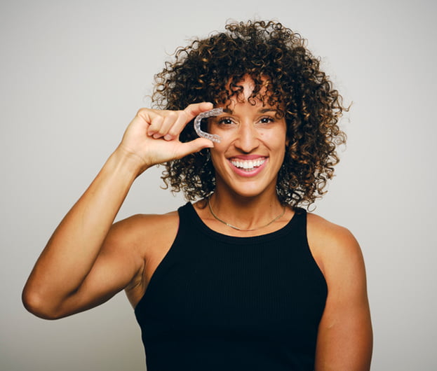A woman with curly hair holding up an Invisalign, smiling and showcasing it in her hand