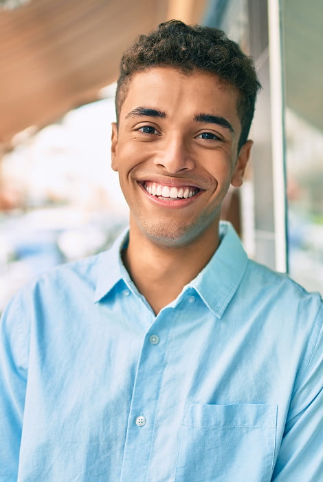 A young man smiles brightly while standing in front of a window, with natural light illuminating his face
