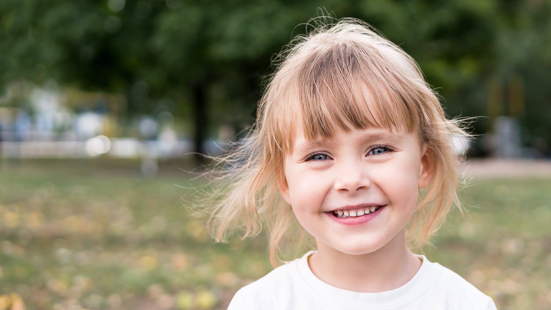 A smiling little girl enjoys a sunny day in the park, surrounded by green grass and trees