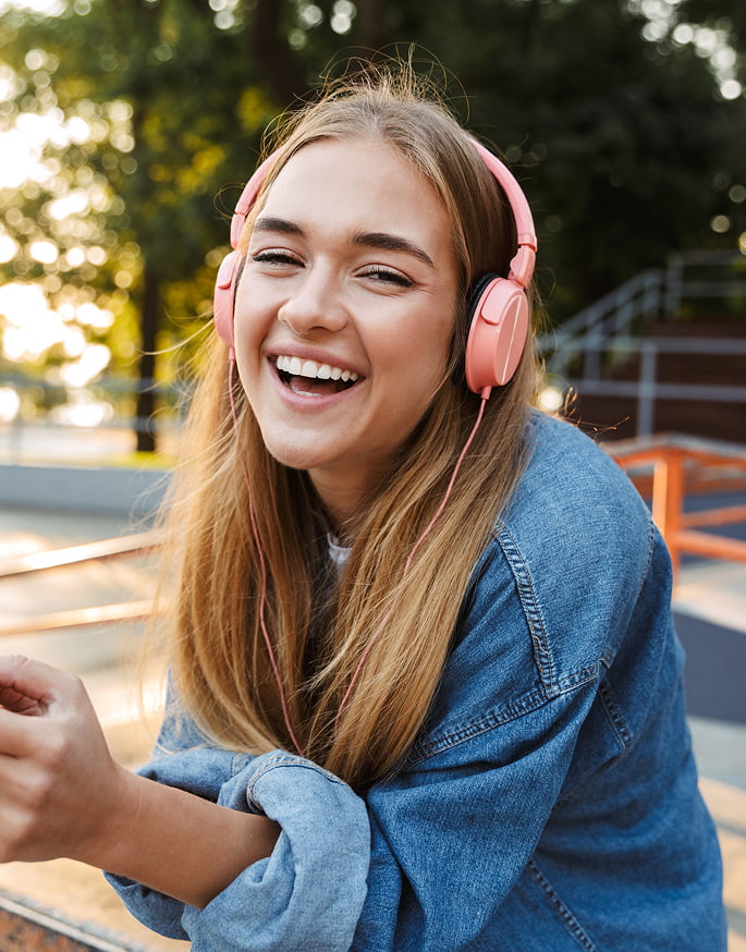A smiling girl wearing headphones sits on a skateboard, enjoying her time outdoors